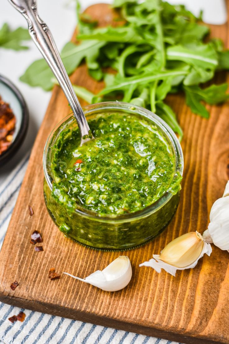 a wooden cutting board topped with a jar of pesto sauce next to garlic and herbs