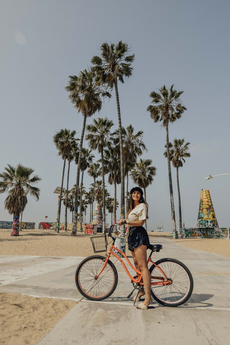 a woman standing next to her bike on the beach with palm trees in the background