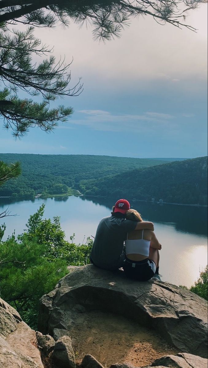 two people sitting on top of a large rock next to a lake in the woods