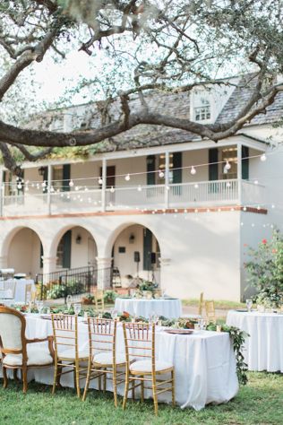 an outdoor table set up with white linens and gold chairs under a large oak tree