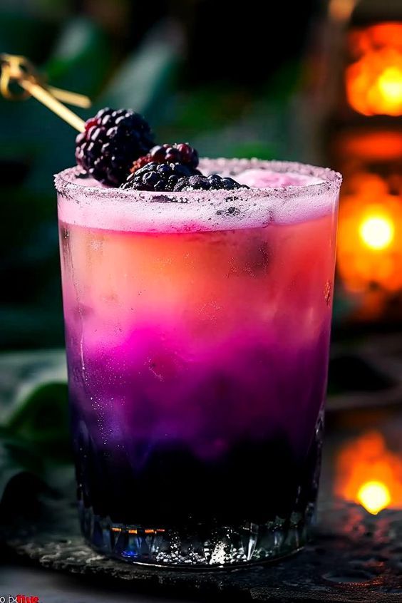 a purple drink with blackberries on the rim and garnished with ice, sits in front of lit candles