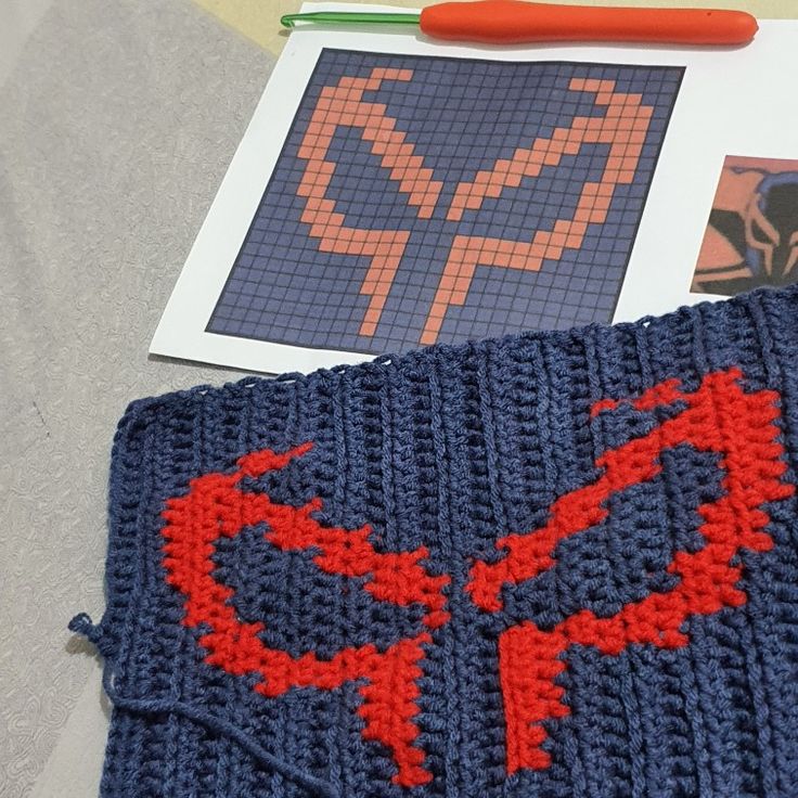 a crocheted spiderman blanket next to an orange and blue pen on a table