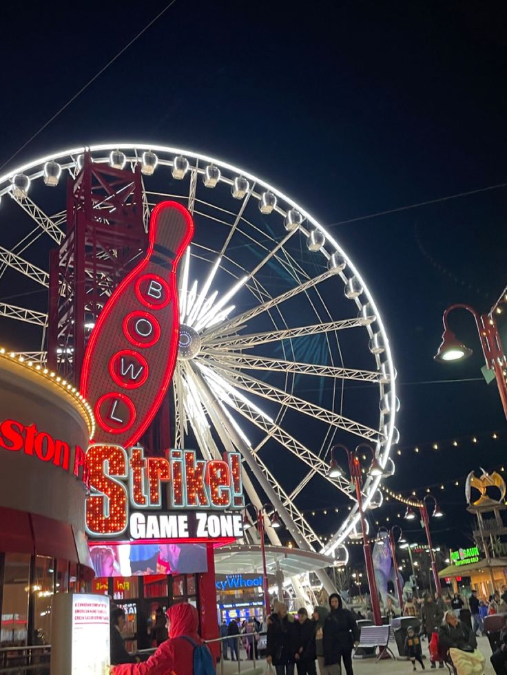 people are walking around in front of a ferris wheel with lights on it at night