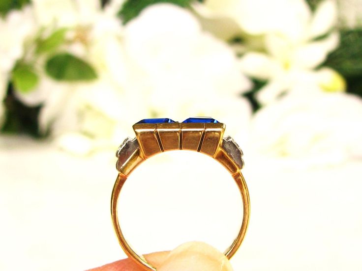 a hand holding a gold ring with two blue stones on it and white flowers in the background