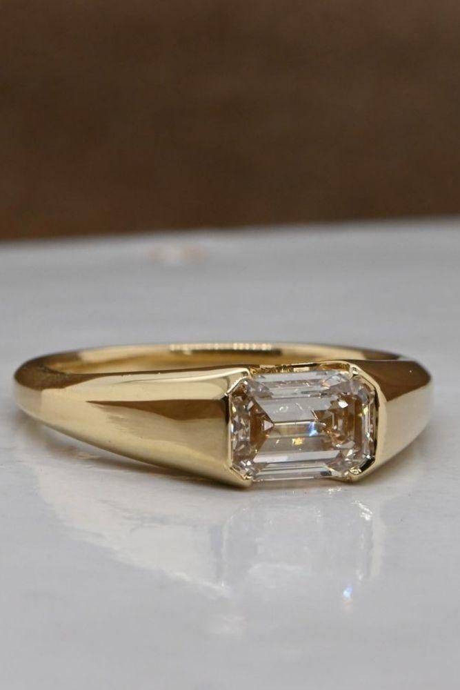 a yellow gold ring with an emerald cut diamond in the center on a white surface