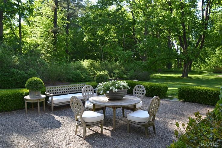 a table and chairs in a graveled area surrounded by hedges, bushes and trees