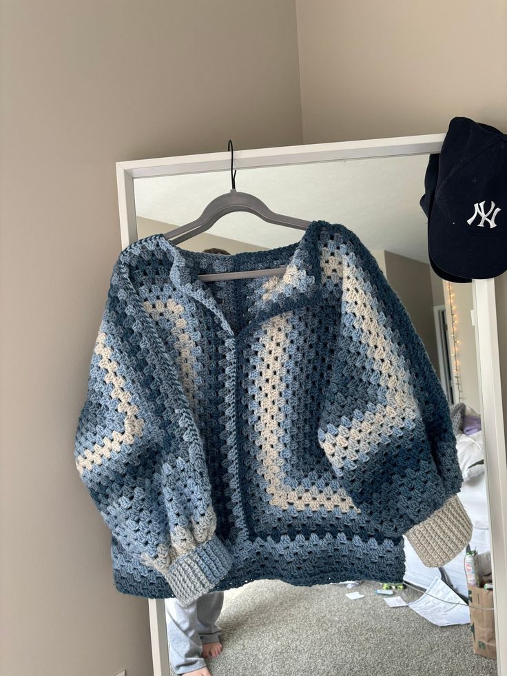 a blue and white crocheted sweater hanging on a mirror next to a hat