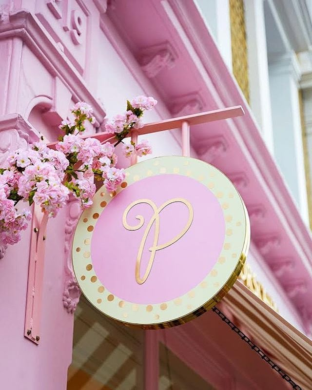 a pink and gold sign hanging from the side of a building with flowers in front of it