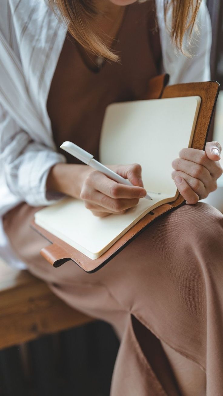 a woman sitting on a bench writing in a note book with a pen and paper