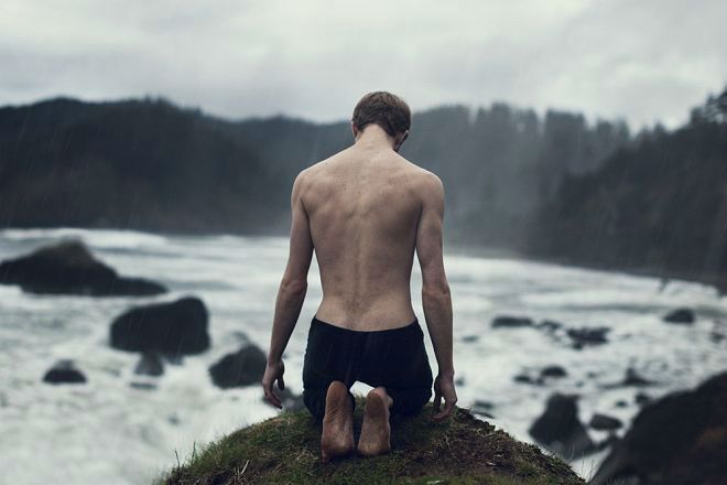 a shirtless man sitting on top of a cliff overlooking the ocean with his feet in the water