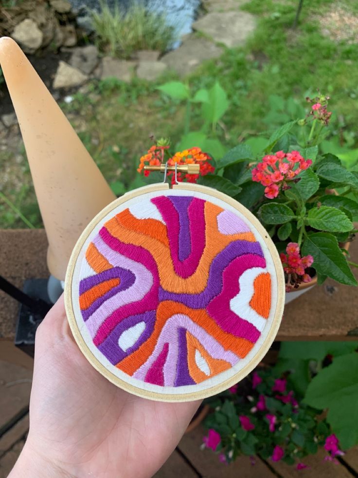 a hand holding up a small colorful embroidered object with flowers in the back ground behind it