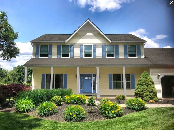 a yellow house with blue shutters and flowers in the front yard on a sunny day