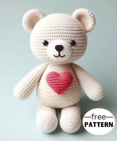 a crocheted teddy bear with a heart on it's chest, sitting against a gray background