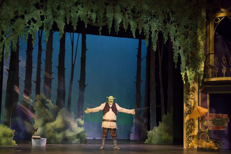 a man in costume standing on stage with his arms spread out to the side and trees around him