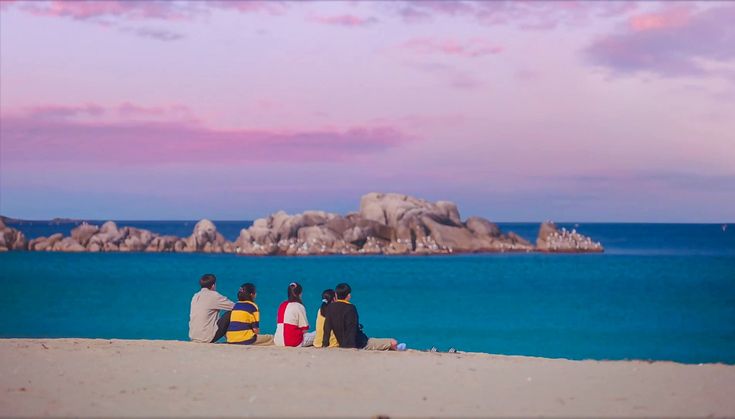 three people sitting on the beach looking out at the water and rocks in the distance