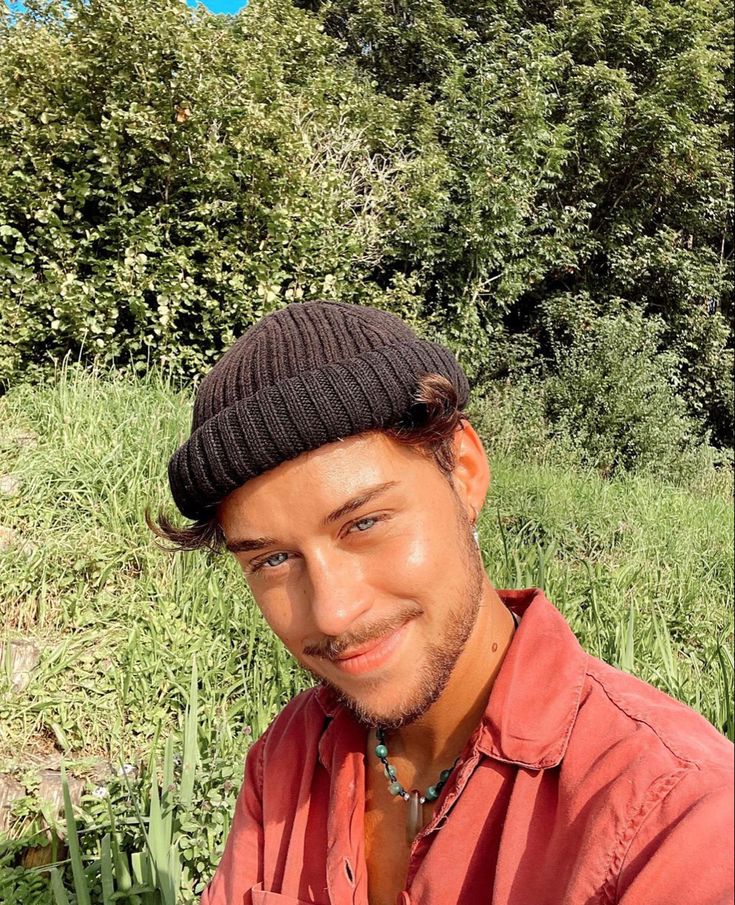 a man in a red shirt and black beanie smiles at the camera with trees behind him