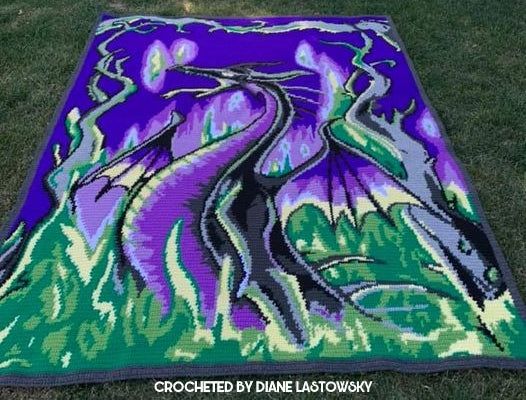 a purple and green blanket on the grass with trees in the background that has been crocheted