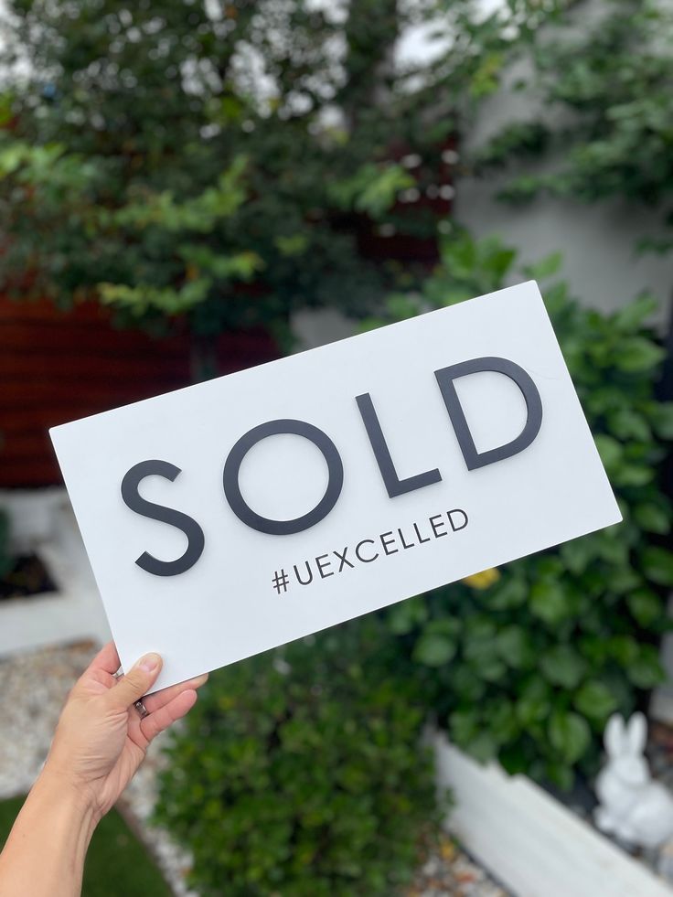 a person holding up a sold sign in front of some plants