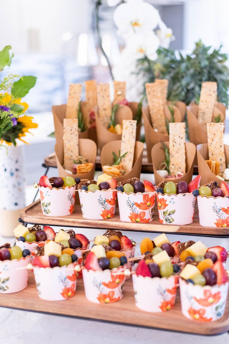 small cups filled with fruit and crackers on top of a wooden tray next to flowers