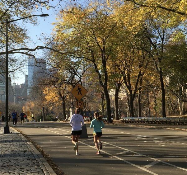 two people are running down the street in front of some trees with yellow and orange leaves