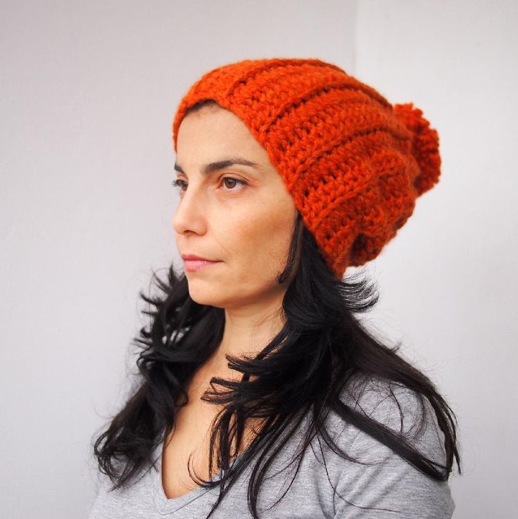 a woman wearing an orange crocheted beanie looks off to the side while standing in front of a white wall