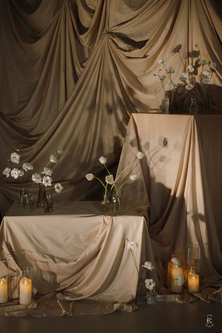 a table topped with lots of white flowers and candles next to a drape covered wall