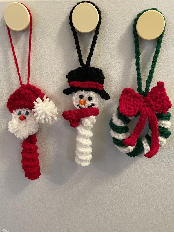 three crocheted snowmen hanging from hooks on a wall, one wearing a santa hat and the other holding a candy cane