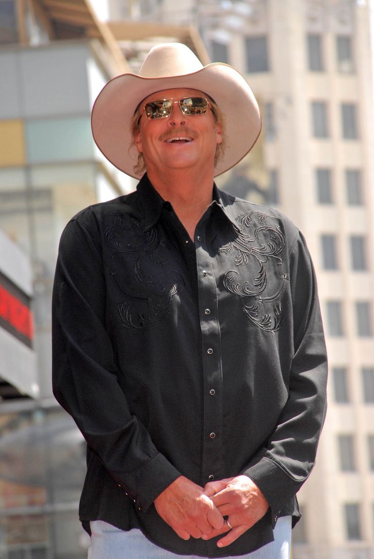 a man wearing a cowboy hat and sunglasses standing in front of a tall building with his hands in his pockets