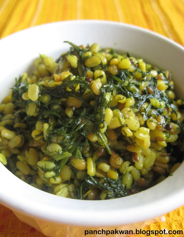 a white bowl filled with green vegetables on top of a yellow table cloth next to a wooden spoon