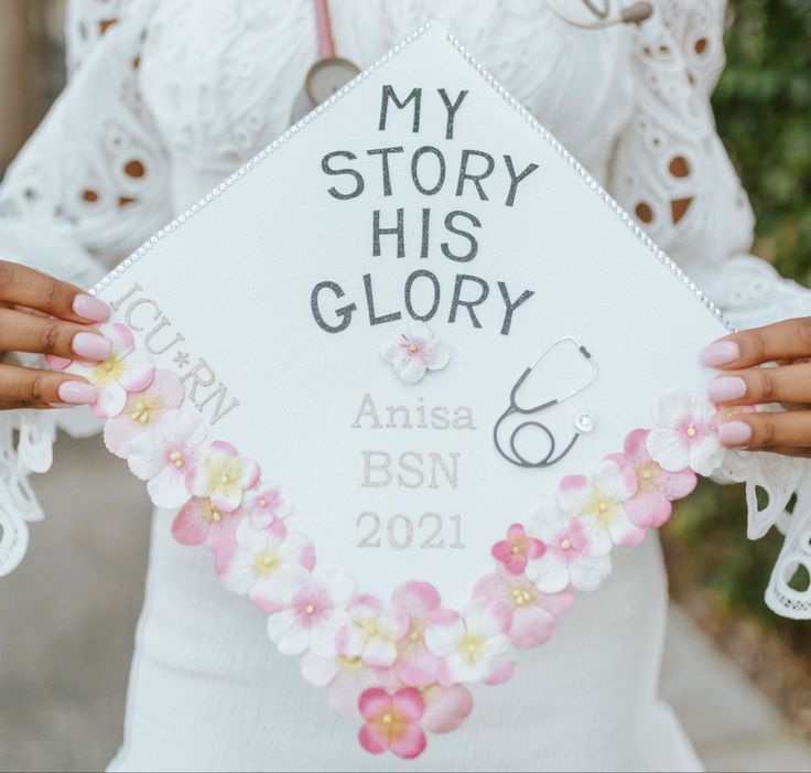 a woman holding up a white graduation cap with flowers on it that reads, my story his glory