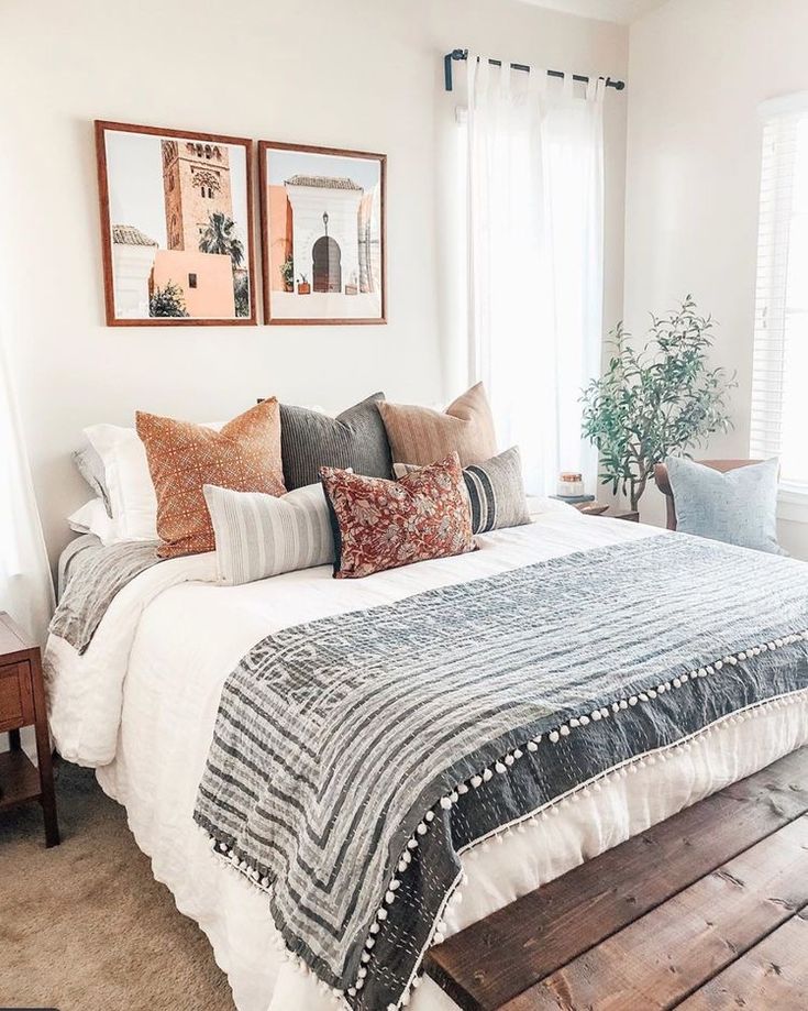 a bed with many pillows on top of it in a bedroom next to two windows