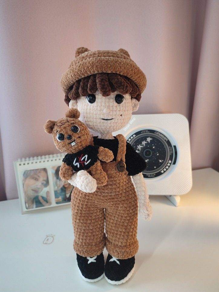 a crocheted doll holding a teddy bear on top of a white table next to a clock