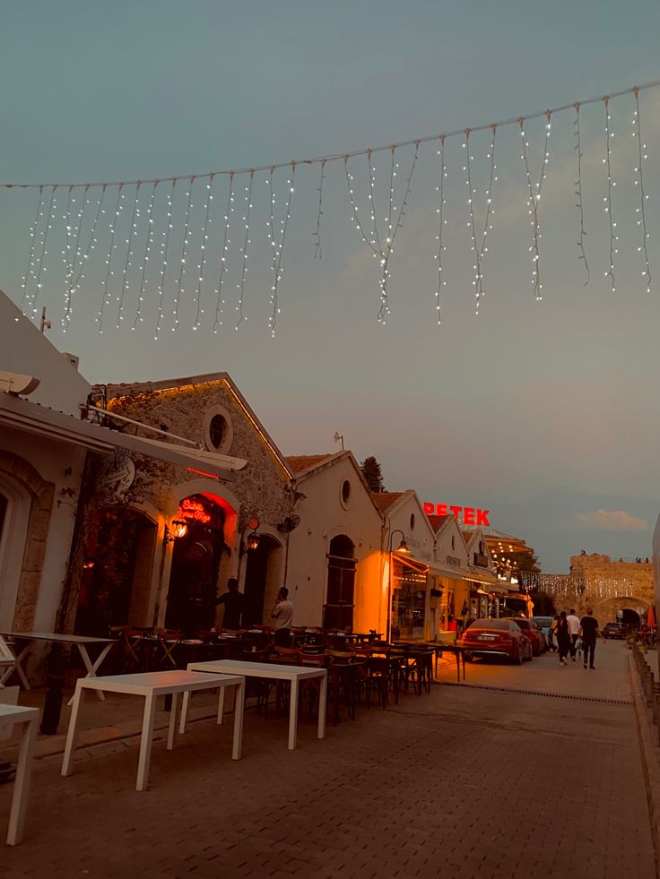 people are walking down the street in front of buildings with lights strung over them and tables on either side