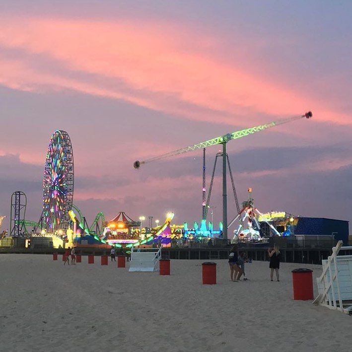 the ferris wheel is lit up at dusk on the beach with people walking around it