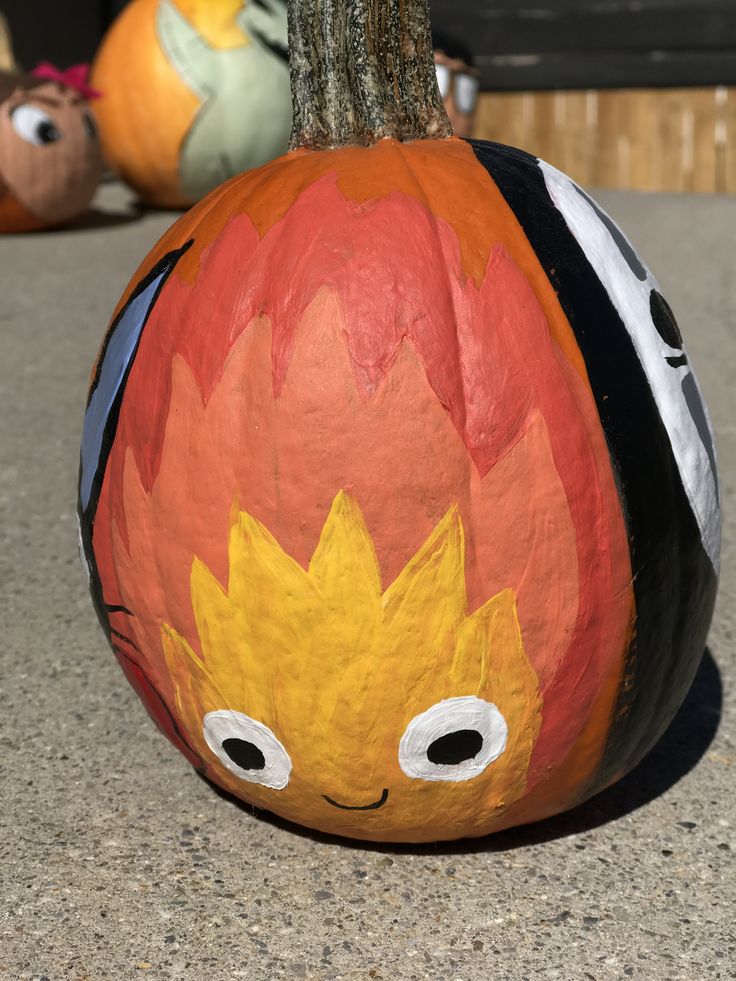 a painted pumpkin sitting on top of a cement ground next to a tree trunk with eyes drawn on it