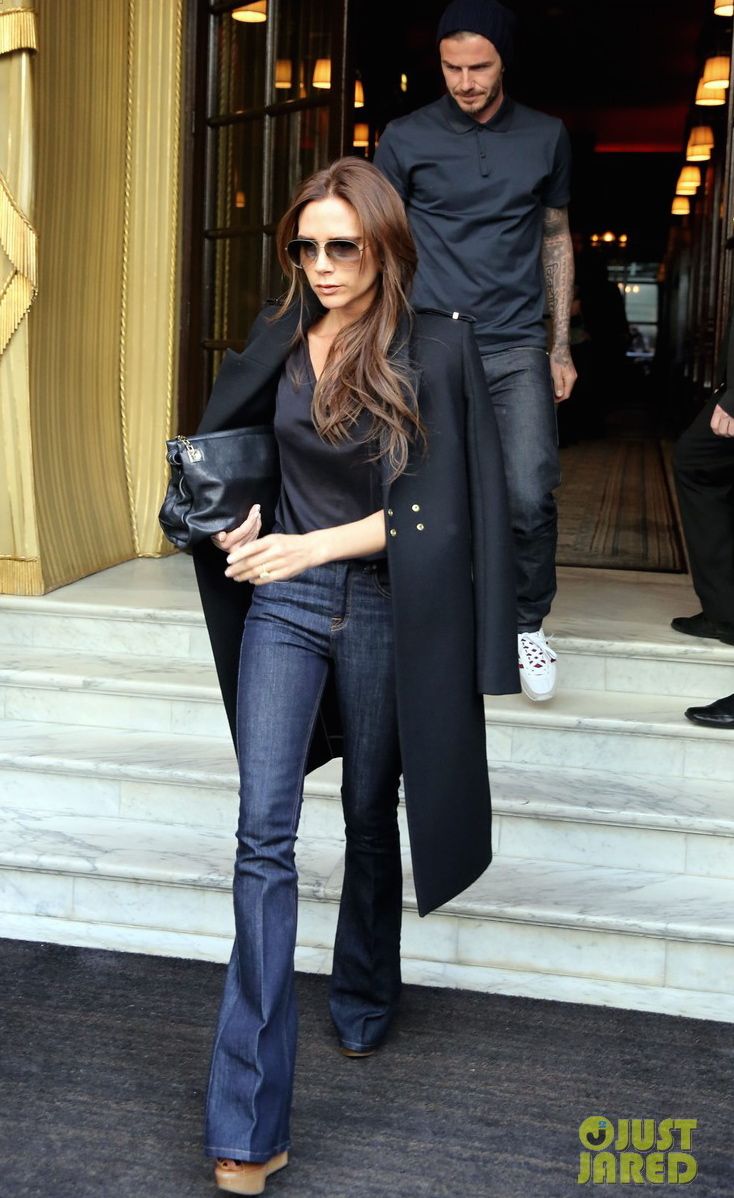 a woman in black coat and jeans walking out of a building with a man standing behind her