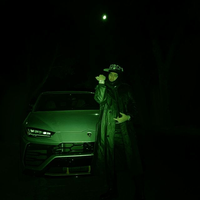 a man standing next to a green car in the dark with his hand on his hip