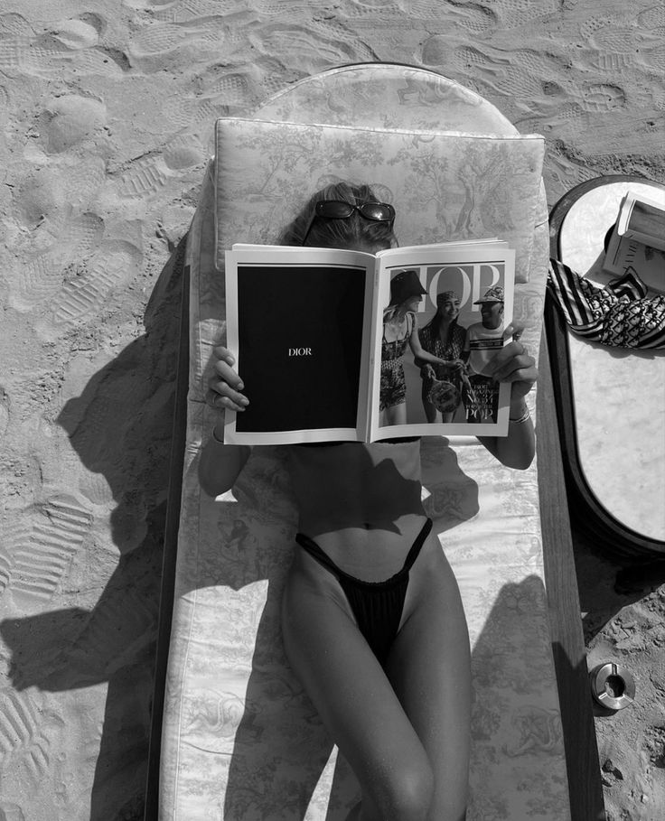 a woman laying on top of a beach next to a mirror holding an open book