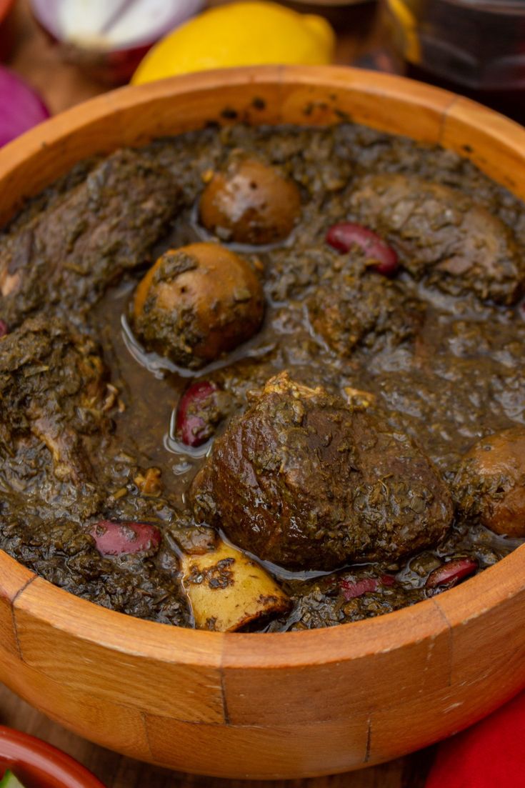 a wooden bowl filled with meat and potatoes on top of a table next to vegetables