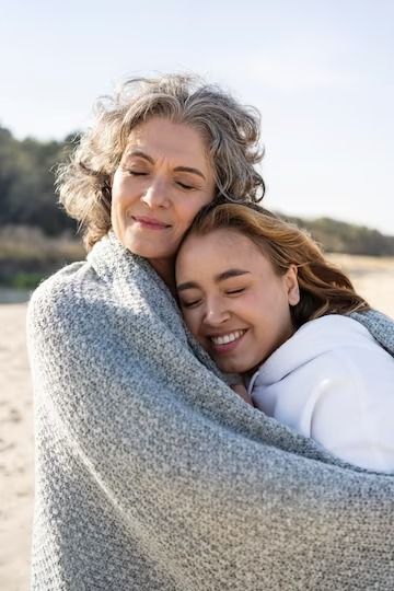 an older woman wrapped in a blanket on the beach hugging her younger daughter's shoulders