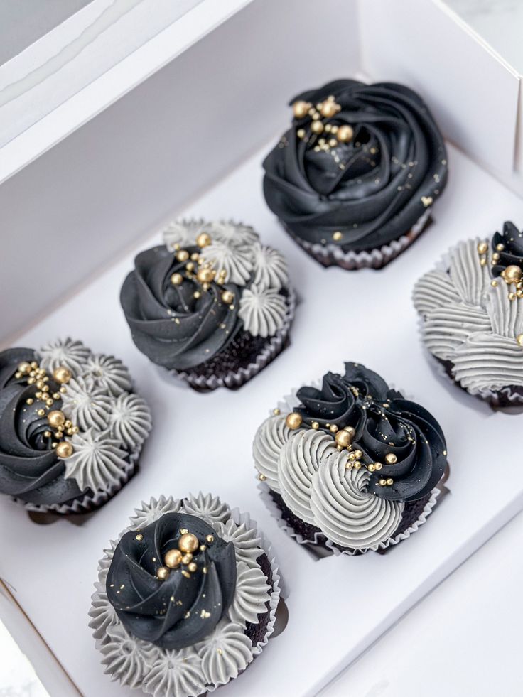 six cupcakes with black frosting and gold sprinkles in a white box