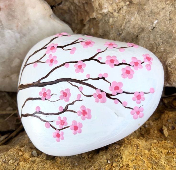 a painted rock sitting on the ground next to a large rock with pink flowers and brown branches