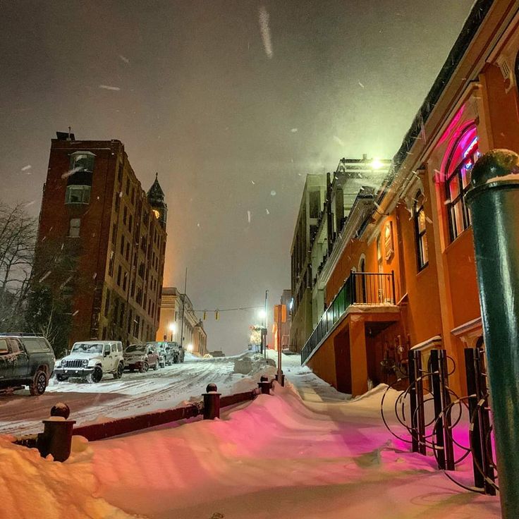 a snowy street with cars parked on the side and buildings in the background at night