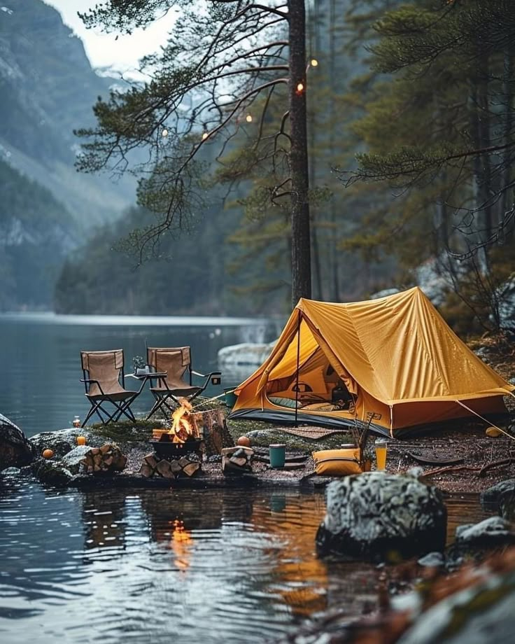 a tent is set up on the shore of a lake with two chairs around it