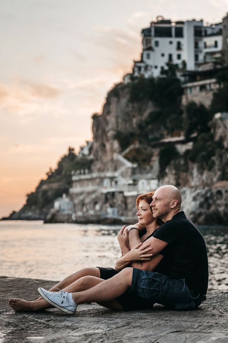 a man and woman sitting next to each other on the beach with buildings in the background