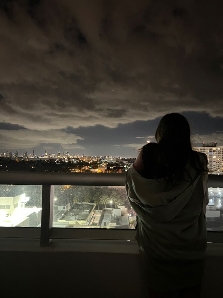 a woman standing in front of a window looking out at the city lights and buildings