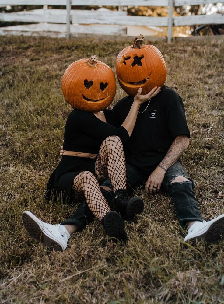 two people sitting in the grass with pumpkins on their heads