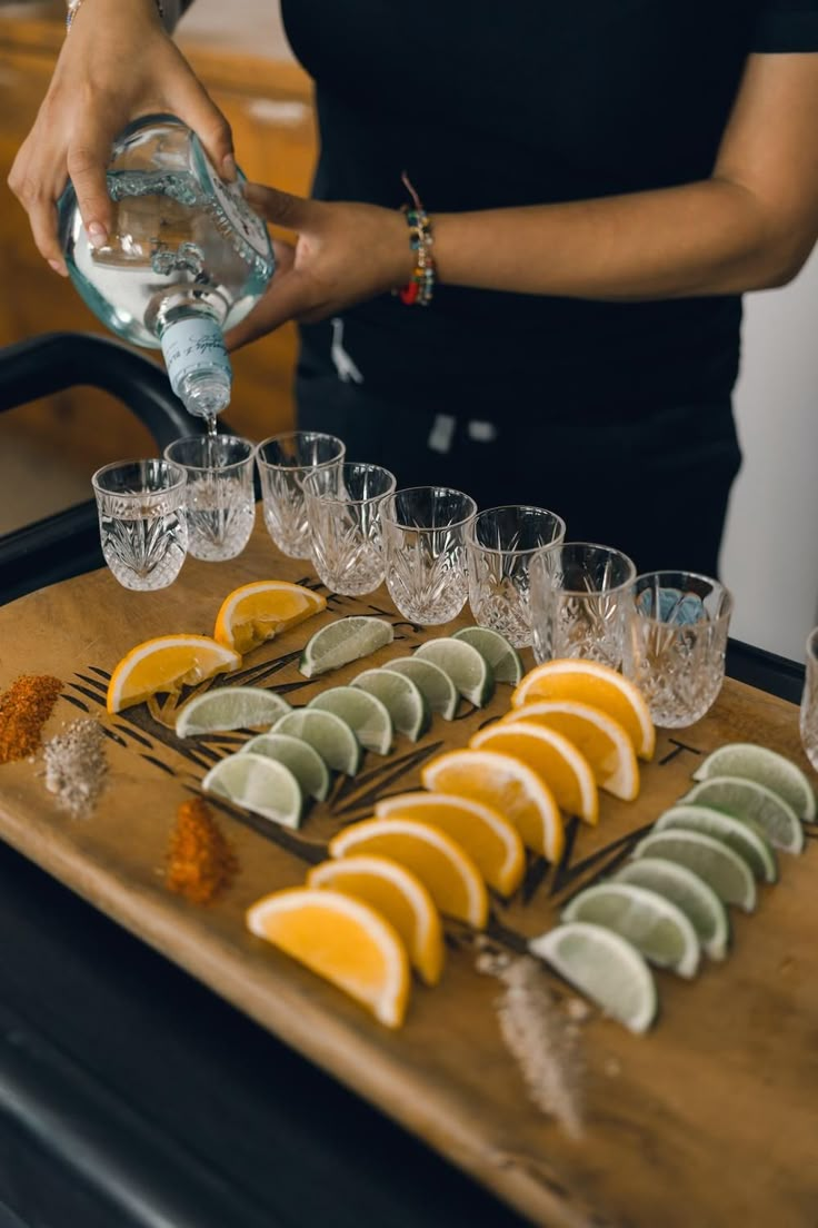 a woman pouring water into glasses on top of a wooden tray filled with lemons and lime slices