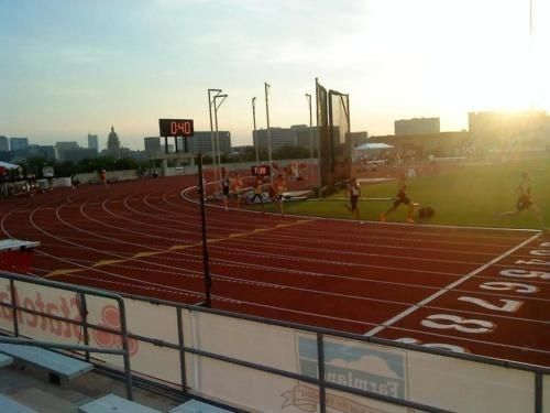 the sun is setting on a track with people running in it and some buildings are in the background