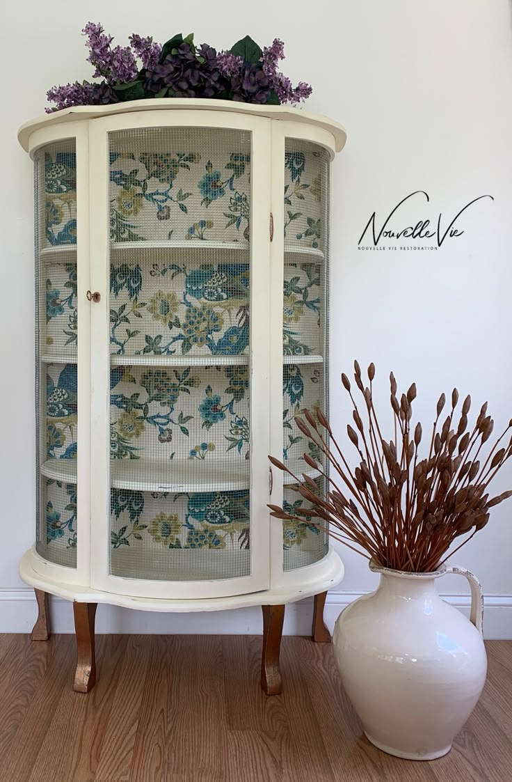 a white china cabinet next to a vase with purple flowers on it and a wallpapered background